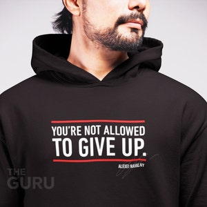 Peut inclure: Sweat-shirt à capuche noir avec le texte "YOU'RE NOT ALLOWED TO GIVE UP." en blanc avec une bordure rouge. Le texte "ALEXEI NAVALNY" est écrit en blanc plus petit sous le texte principal avec une ligne de signature.