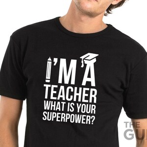 May include: Black t-shirt with white text that reads "I'm a teacher what is your superpower?" with a pencil and graduation cap graphic.