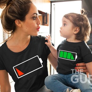 May include: A woman and a child wearing matching black t-shirts with white battery icons. The woman's battery icon is red, indicating low battery, while the child's battery icon is green, indicating full battery.