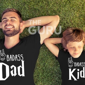 May include: Two black t-shirts with the text "BADASS Dad" and "BADASS Kid" in white, with a hand gesture graphic. The shirts are worn by a man and a child lying on green grass.