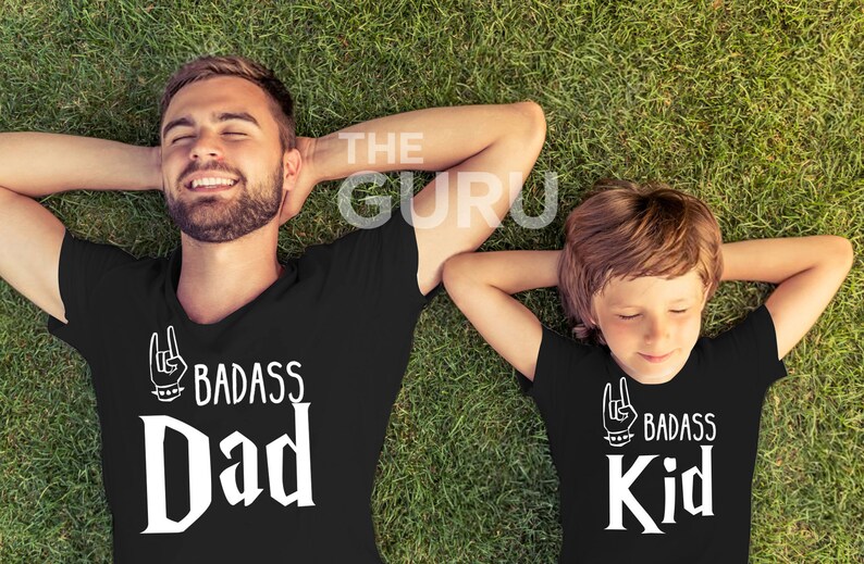 May include: Two black t-shirts with the text "BADASS Dad" and "BADASS Kid" in white, with a hand gesture graphic. The shirts are worn by a man and a child lying on green grass.