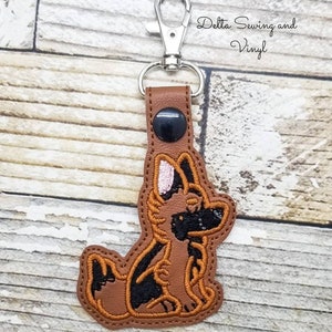 May include: A brown leather keychain with a silver clip and a black button. The keychain features an embroidered image of a German Shepherd dog.