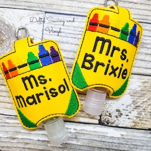 May include: Two yellow hand sanitizer holders with a crayon box design. The holders have a green border and a clear plastic bottle. The text on the holders reads "Mrs. Briskie" and "Mrs. Marisol".