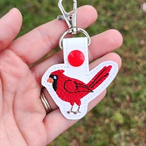 May include: A red cardinal bird keyring with a white backing and a silver clasp. The bird is embroidered with black and red thread, and the keyring has a red button. A decorative accessory.