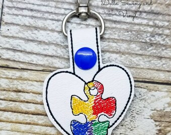 May include: White leather keychain with a blue button and a heart-shaped design. The heart contains a colorful puzzle piece. The text "Delta Sewing and Vinyl" is visible in the background.