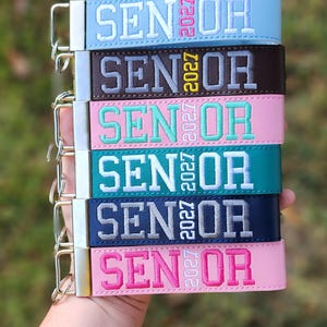 May include: A stack of six rectangular wristlet keychains in light blue, dark brown, light pink, teal, navy blue, and bright pink. Each features the embroidered text SENIOR with the year 2027 vertically placed between the letters.
