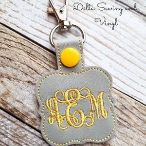 May include: A gray leather keychain with a yellow button and gold embroidered initials "CGM". The keychain is attached to a silver split ring.