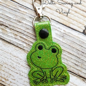 May include: Green glitter frog keychain with black eyes and a black button on the top. The keychain has a silver metal clasp.