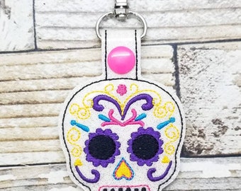 May include: White sugar skull keychain with colorful embroidery details. The skull has black eyes, a yellow heart, and a purple and pink floral design. The keychain has a silver metal clasp and a pink button.