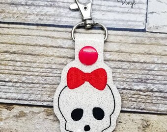 May include: White glitter skull keychain with a red bow and a pink button. The keychain is attached to a silver metal key ring.