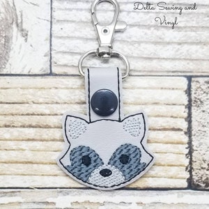 May include: A gray raccoon face keychain with a silver metal key ring. The keychain is made of leather and has a black button on the back.