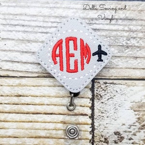 May include: A white leather badge holder with a red monogram "AEM" and a black airplane graphic. The badge holder is attached to a clear retractable badge reel.