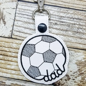May include: White leather keychain with a black embroidered soccer ball and the word "dad" underneath. The keychain has a silver metal clasp.