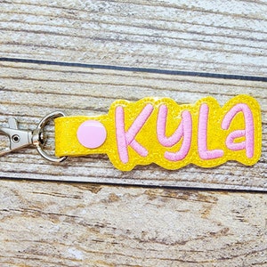 May include: A yellow felt keychain with a pink circle and the name "KYLA" embroidered in pink.