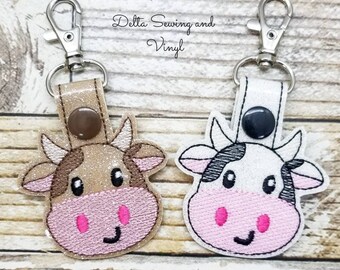 May include: Two cow-themed keychains with a brown cow and a black and white cow. Both keychains have a pink nose and pink cheeks. The keychains are attached to silver key rings.