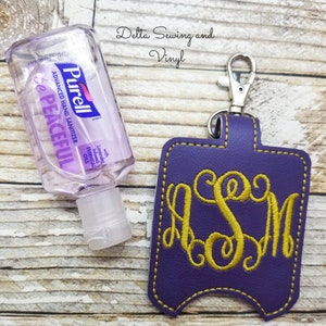 May include: Purple leather hand sanitizer holder with a gold monogram "ASM" and a silver clip. The holder is on a wood background next to a clear bottle of hand sanitizer.