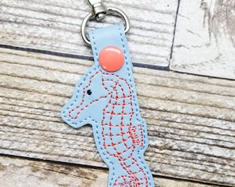 May include: A light blue leather keychain with a red embroidered seahorse design. The keychain has a silver metal lobster clasp and a coral-colored button.