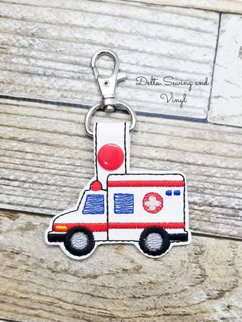 May include: White ambulance keychain with red and blue accents. The keychain features a red cross emblem and black wheels. The text "Delta Sewing and Vinyl" is visible in the background.