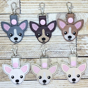 May include: Six keychains featuring embroidered chihuahua dog faces in various colors, including brown, gray, white, and tan. Each keychain has a silver metal ring and a pink button.