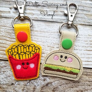 May include: Two keychains with a cute food theme. One keychain features a red and yellow cartoon french fry container with a smiling face. The other keychain features a tan cartoon hamburger with a smiling face. Both keychains have a silver metal clasp.