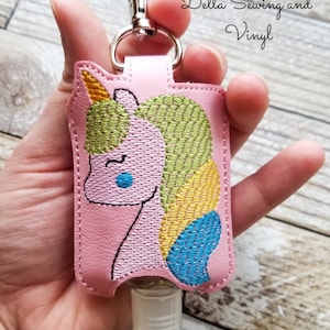 May include: Pink hand sanitizer holder with a unicorn design embroidered in green, yellow, and blue thread. The holder has a clear plastic bottle attached and a silver keychain.