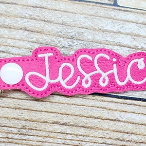 May include: Pink fabric keychain with a white embroidered name "Jessica" and a silver metal clasp.