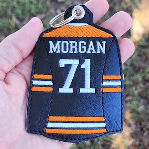 Peut inclure: Porte-clés en forme de maillot noir et orange avec le nom "MORGAN" et le numéro "71" en blanc. Le design comprend des rayures orange et blanches. Il est équipé d'un anneau métallique pour attacher les clés.