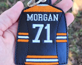 May include: Black and orange jersey-shaped keychain with the name "MORGAN" and the number "71" in white. The keychain has a metal ring at the top for attachment. The jersey design includes orange and white stripes.