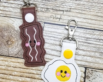 May include: Two keychains, one shaped like a piece of bacon and the other shaped like a fried egg. Both keychains have a smiling face with pink cheeks. The bacon keychain is brown with white stripes and the egg keychain is white with a yellow yolk.