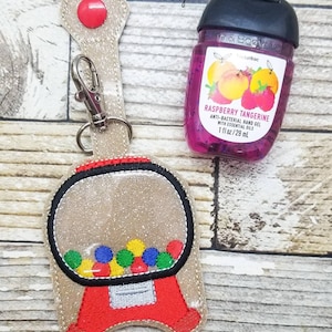 May include: A beige gumball machine keychain with a clear plastic window and colorful gumballs inside. The keychain is attached to a silver clip. A small bottle of raspberry tangerine hand sanitizer is also pictured.