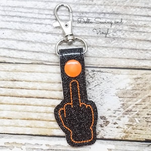 May include: Black glitter keychain with an orange middle finger and a silver key ring. The keychain has the text "Delta Sewing and Vinyl" on it.