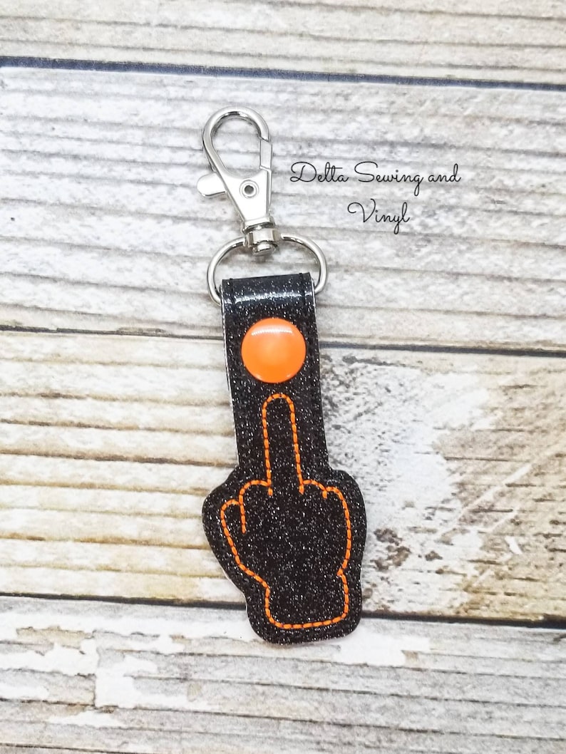 May include: Black glitter keychain with an orange middle finger and a silver key ring. The keychain has the text "Delta Sewing and Vinyl" on it.