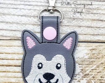 May include: Gray felt husky dog keychain with pink ears and a pink button. The keychain has a silver metal clip.