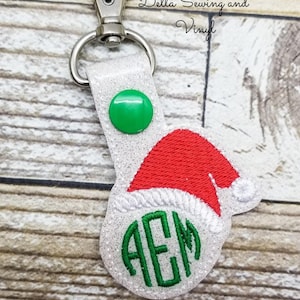 May include: White glitter keychain with a red Santa hat and green embroidered monogram "AEM".