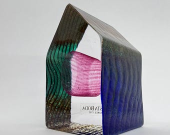 May include: A house-shaped glass sculpture featuring a textured exterior with vertical wavy lines in shades of green and blue. A pink cylindrical glass element is suspended in the centre. The base includes the text KOSTA BODA 1742.