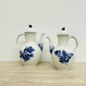 Op de afbeelding: Twee witte porseleinen theepotten met blauwe bloemmotieven. Elke theepot heeft een gebogen handvat, een lange tuit en een deksel met een kleine blauwe knop. Ze staan op een licht oppervlak tegen een witte achtergrond.