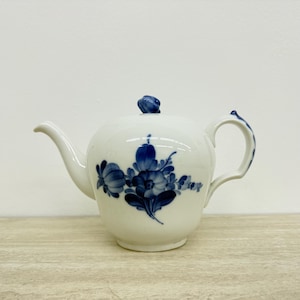 Op de afbeelding: Witte porseleinen theepot met een blauw bloemmotief. De theepot heeft een gebogen tuit, een ronde body en een handvat. Het deksel is bekroond met een kleine blauwe knop. Het bloemmotief bevat gedetailleerde blauwe bloemen en bladeren.
