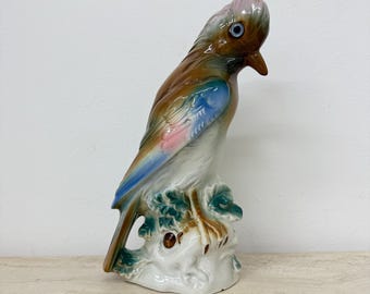 May include: A ceramic bird figurine with a brown body, blue and pink wings, and a pink crest. The bird is perched on a white base with green foliage. The figurine has a glossy finish and is set against a neutral background.