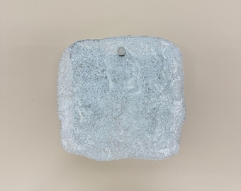 May include: A square-shaped glass object with a textured, frosted, and translucent surface that mimics the appearance of crushed ice. The piece features irregular, jagged edges and a single small, circular metallic stud centered near the top edge.