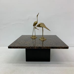 Pode incluir: Uma mesa de centro quadrada de granito com uma base preta. Duas esculturas de pássaros de metal dourado estão sobre a mesa.