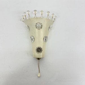 Op de afbeelding: Crèmekleurige wandlamp met een uitlopende vorm, versierd met heldere kristallen bloemvormige accenten en kleine, ronde kristallen versieringen. De lamp heeft een treksnoer en is op een witte muur gemonteerd.