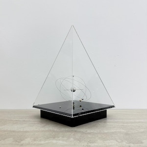 May include: A clear acrylic pyramid display case containing a kinetic orbital sculpture. The sculpture features a central silver sphere surrounded by thin, intersecting metal rings and smaller metallic spheres, all mounted on a black square base.