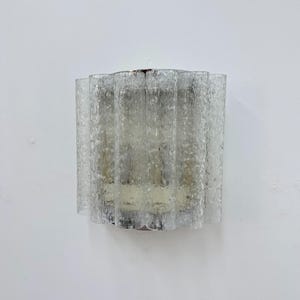 Könnte beinhalten: Eine Wandleuchte bestehend aus einer Reihe vertikaler, klarer Glaspaneele mit einer texturierten, blasigen Oberfläche. Die Glaselemente sind in einem geschwungenen, geriffelten Muster angeordnet und verdecken teilweise die innere Lampenfassung.