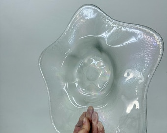 Könnte beinhalten: Eine klare, irisierende Glasschale in Sternform. Die Schale hat eine strukturierte Oberfläche und eine zentrale, abgerundete Vertiefung. Die Ränder sind sanft gewellt, und das Glas reflektiert das Licht mit einem subtilen Regenbogen-Schimmer. Die Schale wird von einer Hand gehalten.