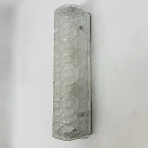 May include: A vertical wall sconce featuring a cylindrical, textured clear glass shade with a repeating geometric pattern. The glass is mounted on a metallic base, secured by two visible screws on the front, creating a light fixture design.