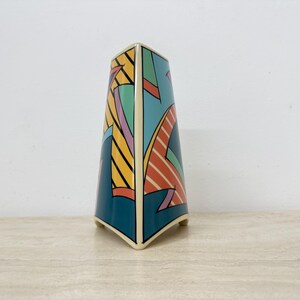 Könnte beinhalten: Eine dekorative Keramikvase mit geometrischem Design. Die Vase hat einen tealfarbenen Sockel mit bunten abstrakten Formen in Gelb, Orange, Rosa und Grün. Die Vase hat eine einzigartige, eckige Form und ist etwa 20 cm hoch.
