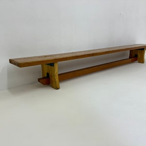 May include: A long wooden bench with a simple design. The bench is made of natural wood with a light brown finish. The bench has a rectangular top and two legs on each end. The legs are angled slightly inward.