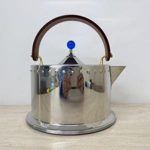 May include: A shiny, stainless steel kettle with a wooden handle and a blue knob on the lid. The kettle has a spout and sits on a round base. The handle is brown and the accents are gold.