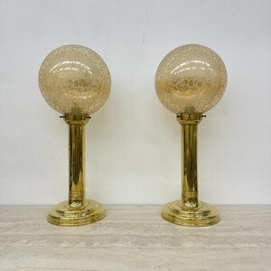 Op de afbeelding: Twee vintage tafellampen. Elke lamp heeft een bolvormige, getextureerde, amberkleurige glazen kap op een messingkleurige, cilindrische steel en een cirkelvormige voet. De lampen worden getoond op een licht oppervlak tegen een witte achtergrond.