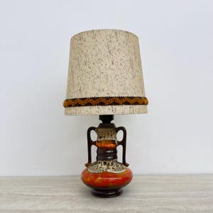 Op de afbeelding: Een tafellamp met een beige lampenkap met textuur en een bruine zigzagrand. De voet is een keramische vaas met twee oren, afgewerkt in een glanzend glazuur van donkerbruin, feloranje en crème met een dikke, druipende lava-achtige textuur.